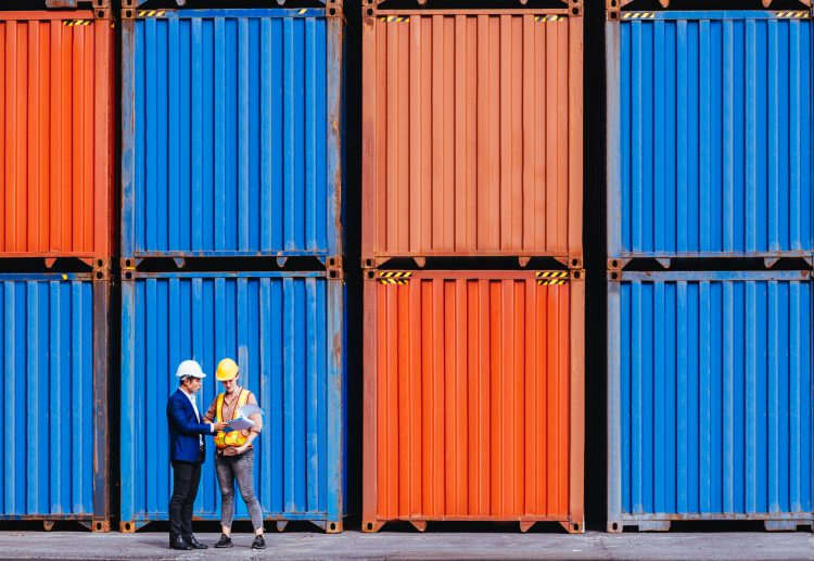 Two men in hard hats stand next to a wall of stacked shipping containers